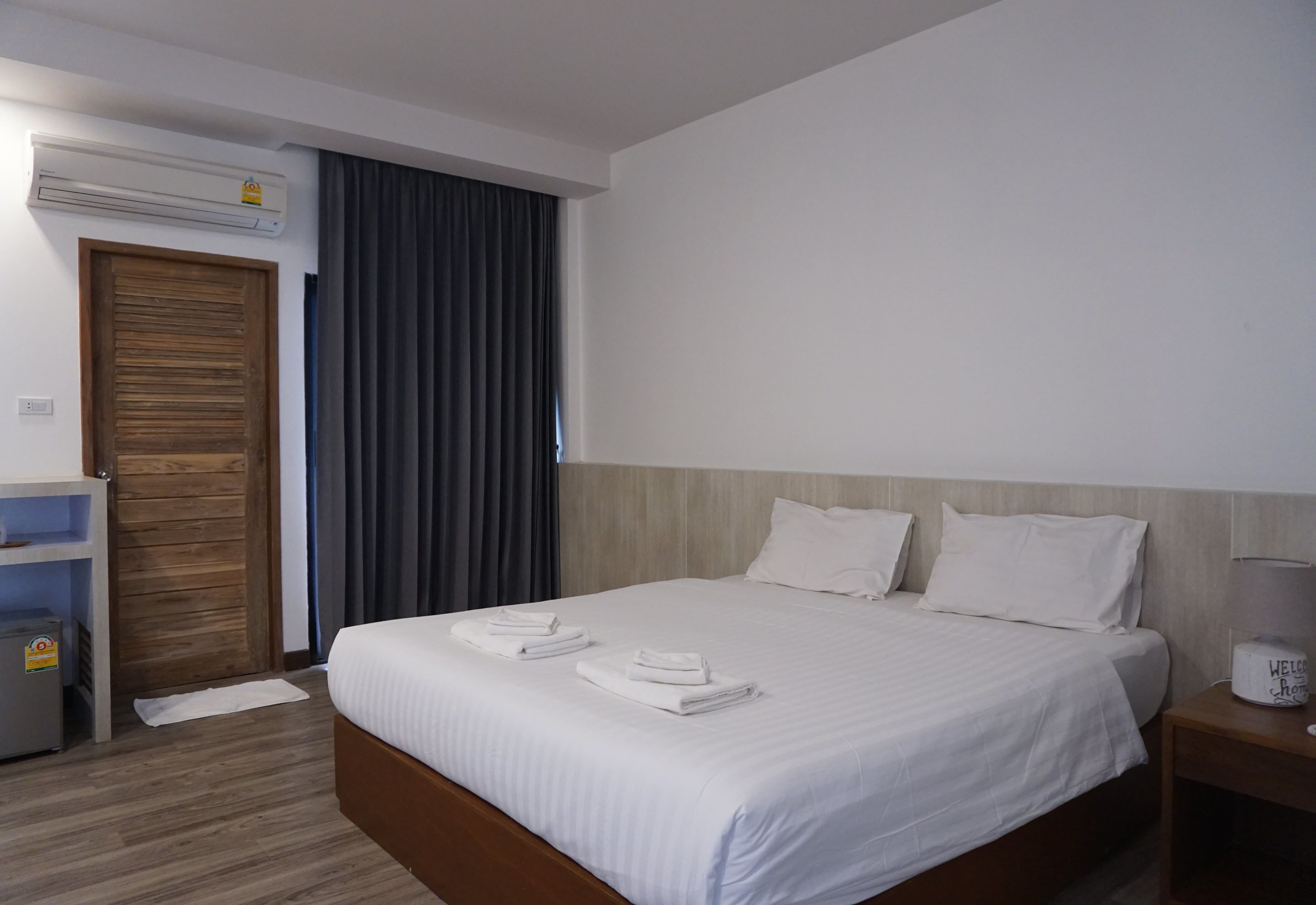 A hotel room with a bed, a wooden door, an air conditioning unit, a nightstand with a lamp, and a towel rack with towels.
