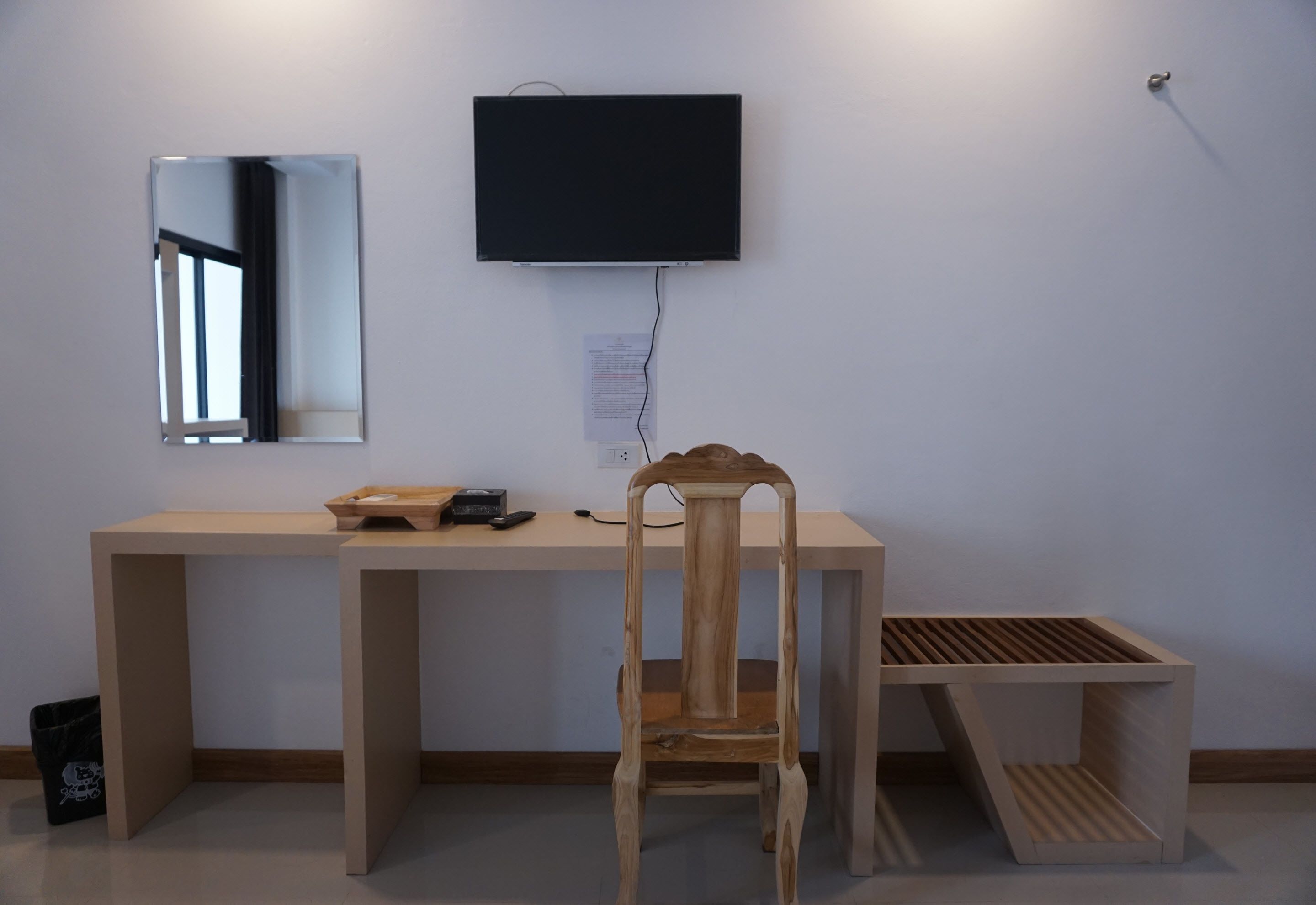 A minimalist office space with a wooden desk, a chair, a television mounted on the wall, and a small trash bin.