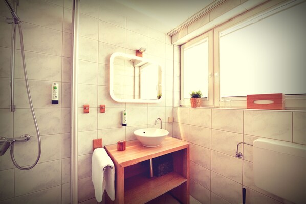 A bathroom with a wooden vanity, a white sink, a mirror, a towel, a shower, and a window. - Melbo’s Home (Lindau)