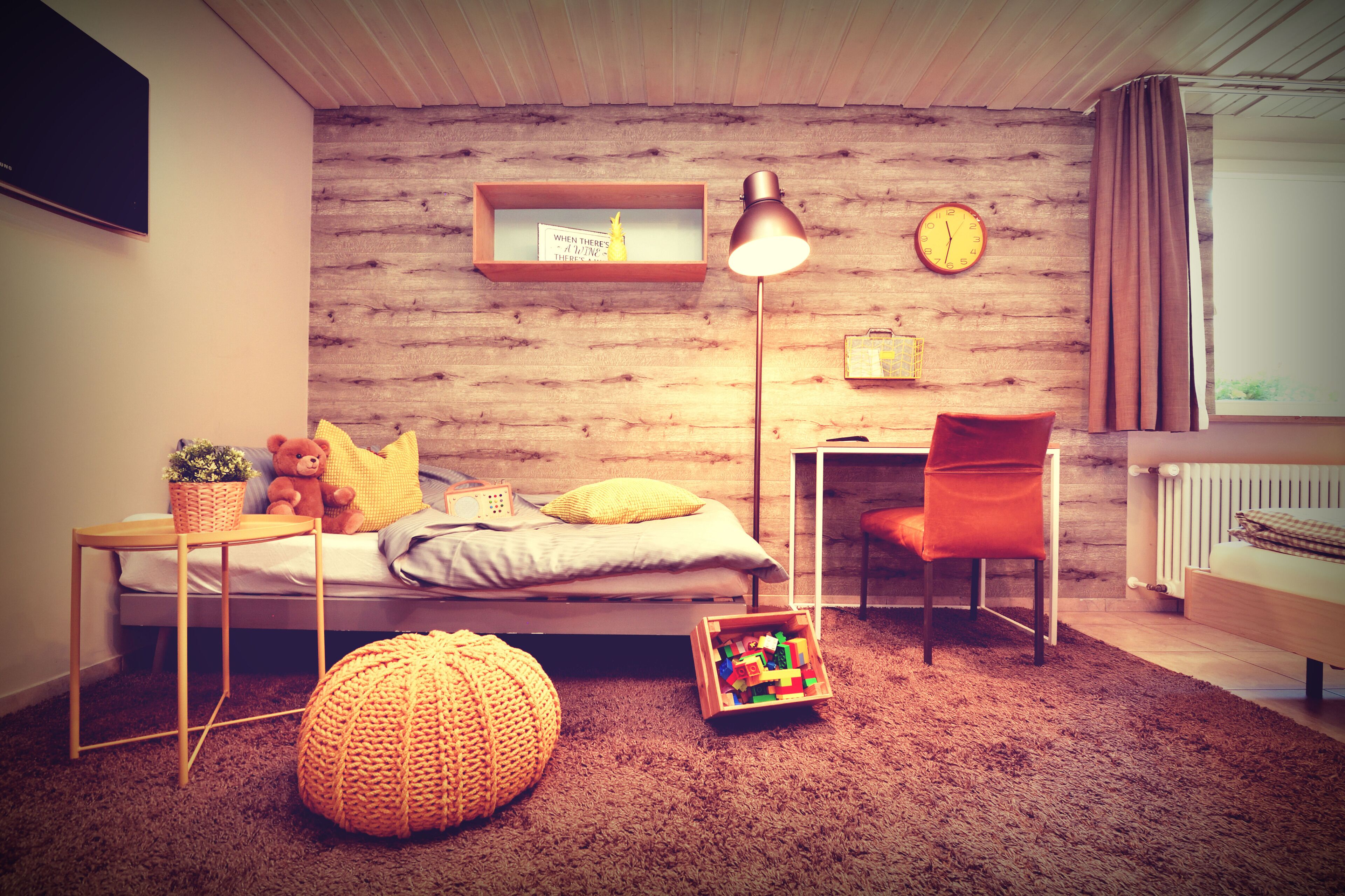 Een knus slaapkamer met een houten wand, een bed met kussens, een bureau, een stoel, een lamp en een klok.