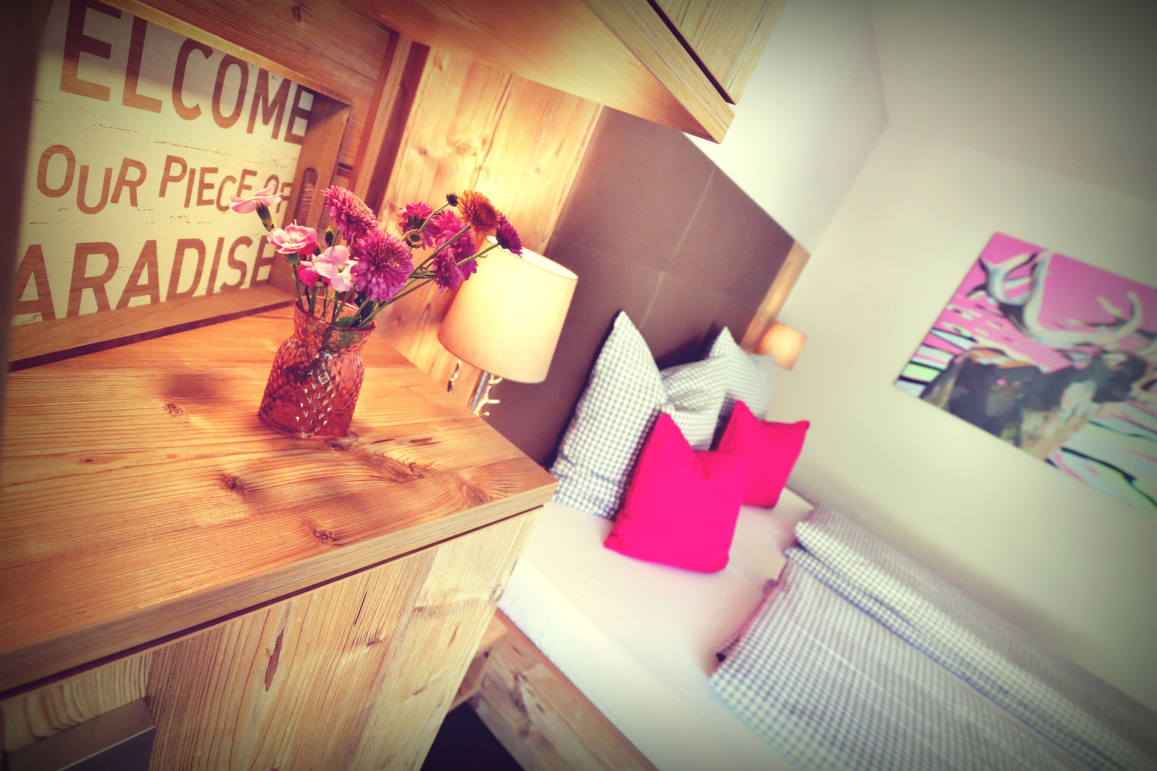 A wooden table with a vase of flowers, a lamp, and a bed with pillows and a blanket.