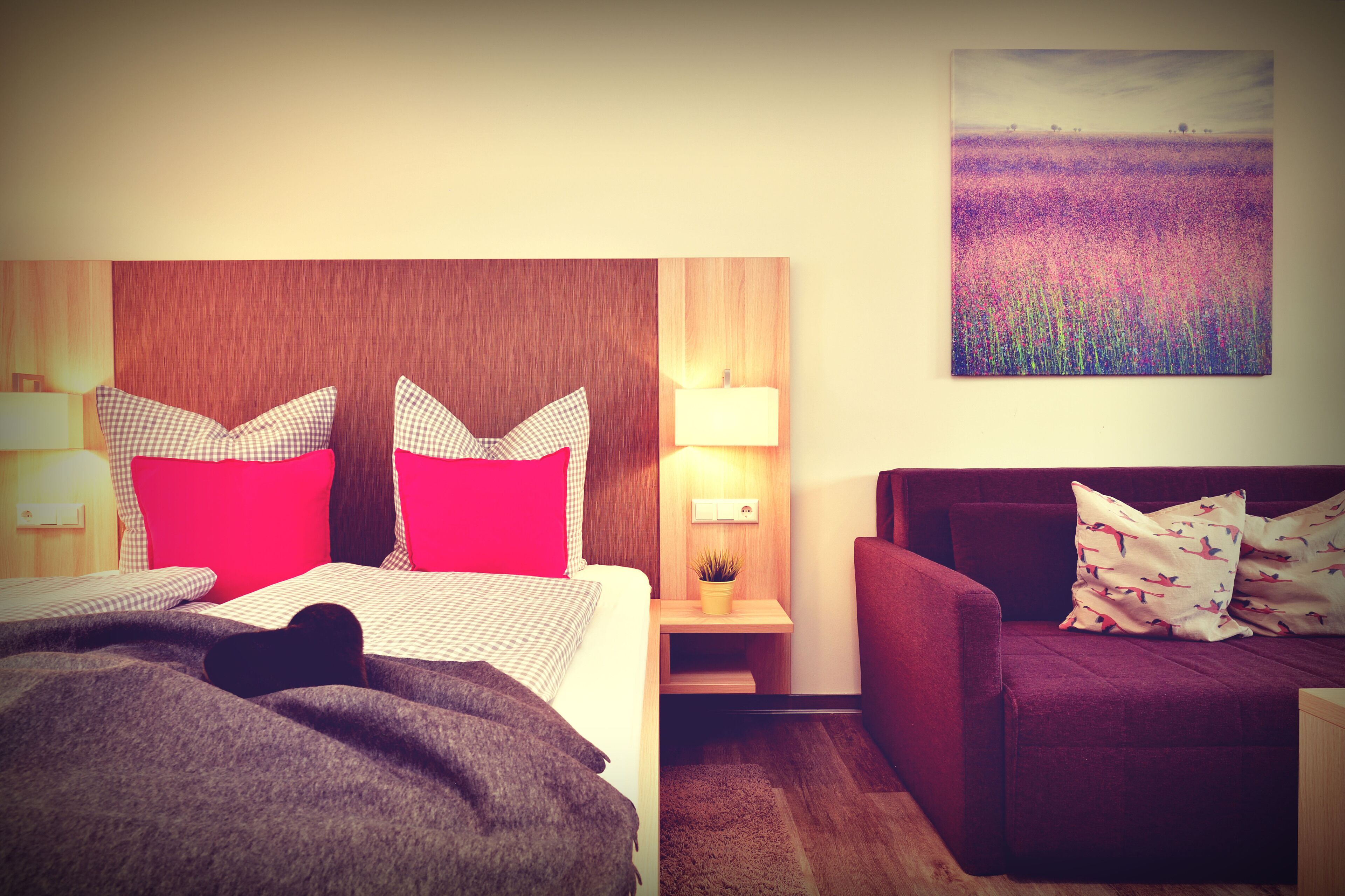 A hotel room with a bed, a sofa, a nightstand with a lamp, and a wall-mounted painting.