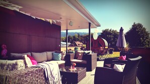 An outdoor seating area with wicker furniture, a glass coffee table, and a view of a house and trees in the background. - Melbo’s Home (Lindau)