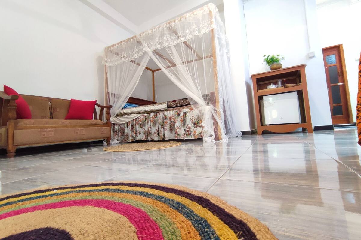 A room with a wooden sofa, a colorful striped rug, a canopy bed, and a small wooden cabinet with a plant.
