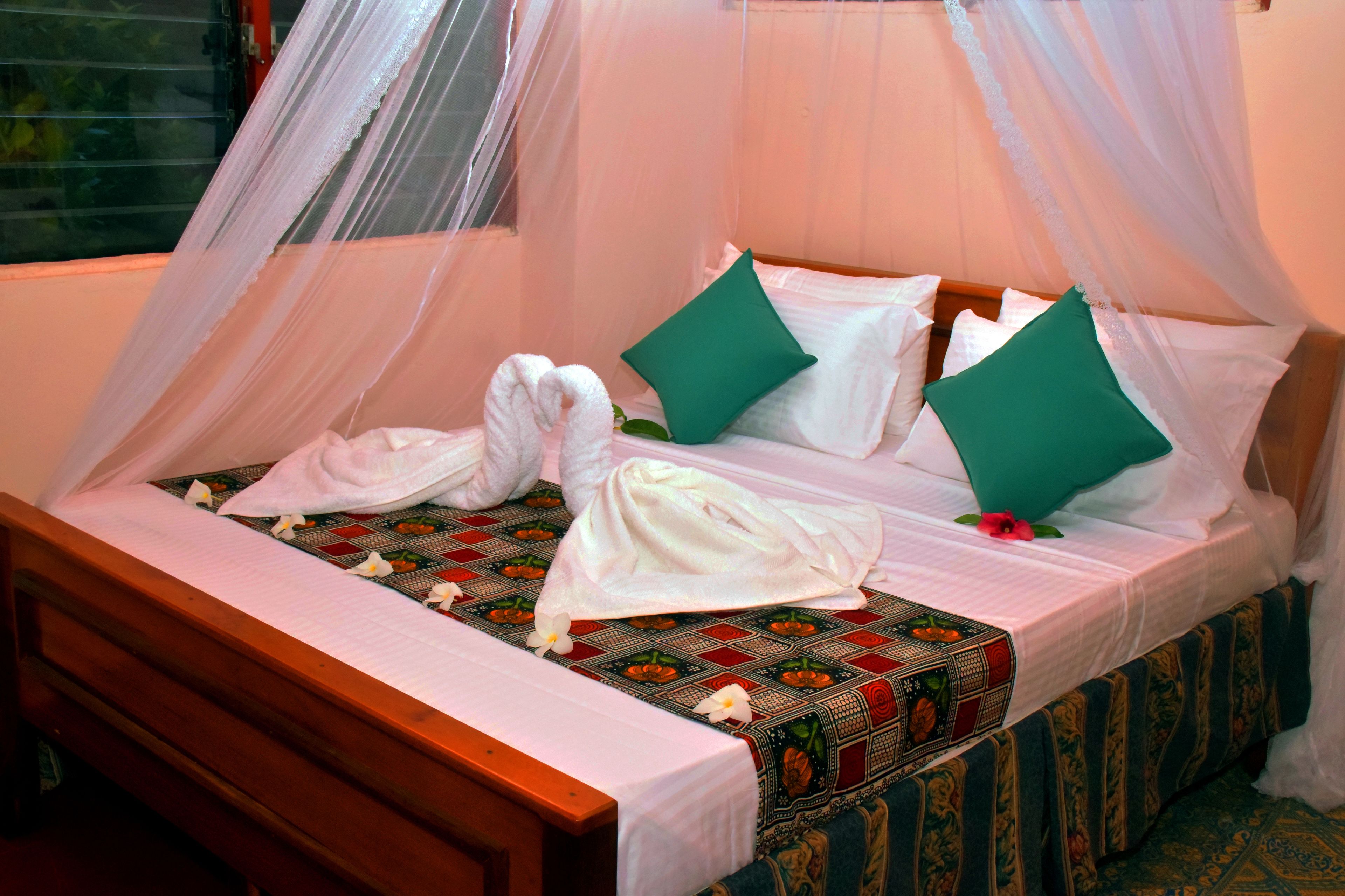 Een bed met hemelbed, witte lakens, groene kussens en een bedsprei met patroon.