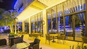 An outdoor seating area with wicker chairs and tables, decorated with string lights, and large windows overlooking a parking area. - Sanya Serenity Coast Marina Hotel (Sanya)