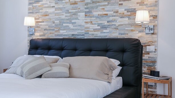 A modern bedroom with a stone-patterned wall, a black upholstered headboard, and two bedside lamps. - Pandolfini Suite (Florence)