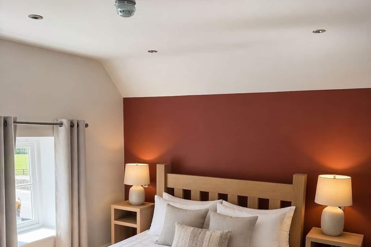 Hotellihuone, jossa on suuri sänky, kaksi yöpöytää valaisimineen, ikkuna verhoineen ja yksi punaisella värillä korostettu seinä.