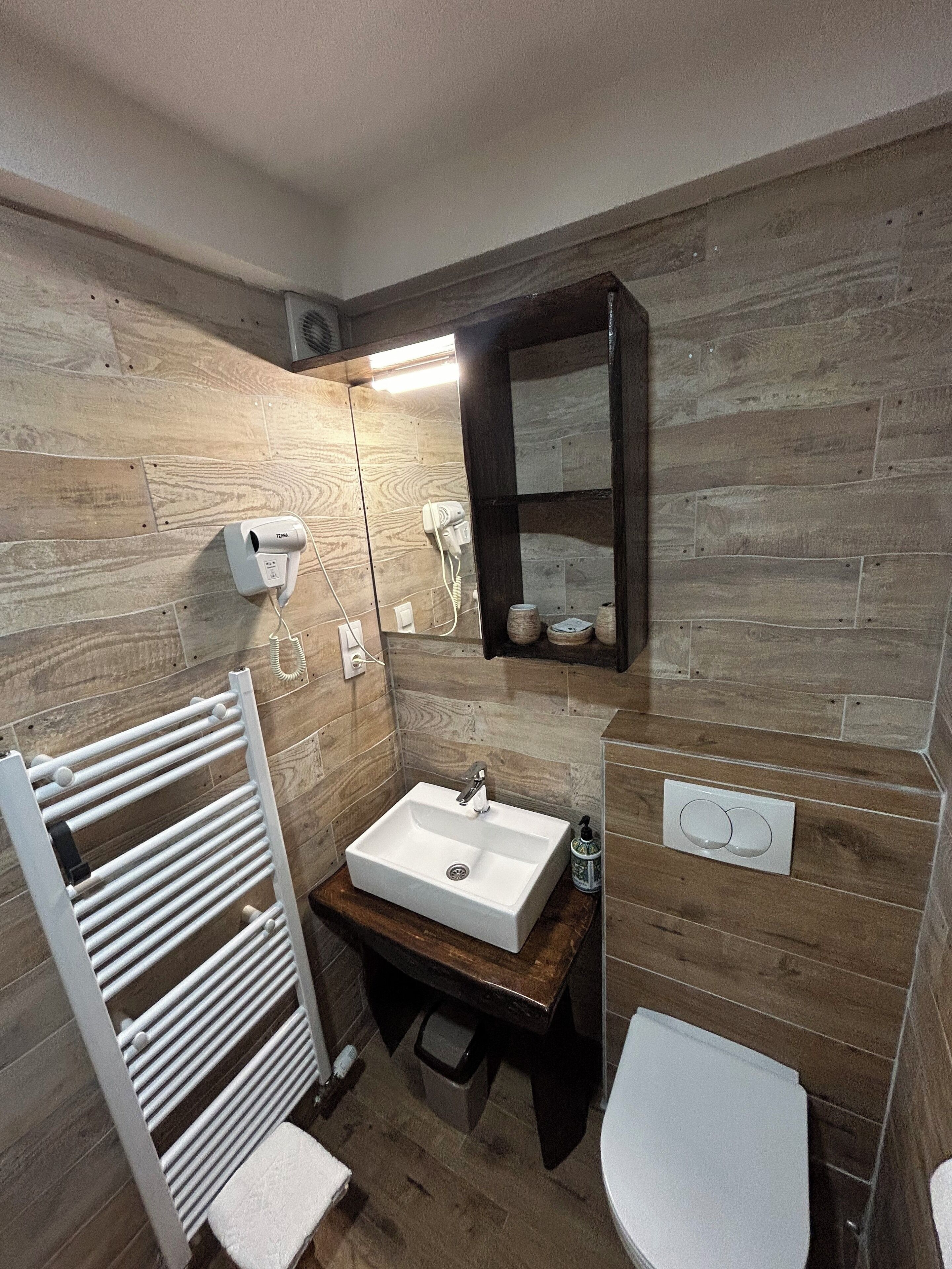 A compact bathroom with a wooden wall, a white sink, a toilet, a towel rack, and a mirror.
