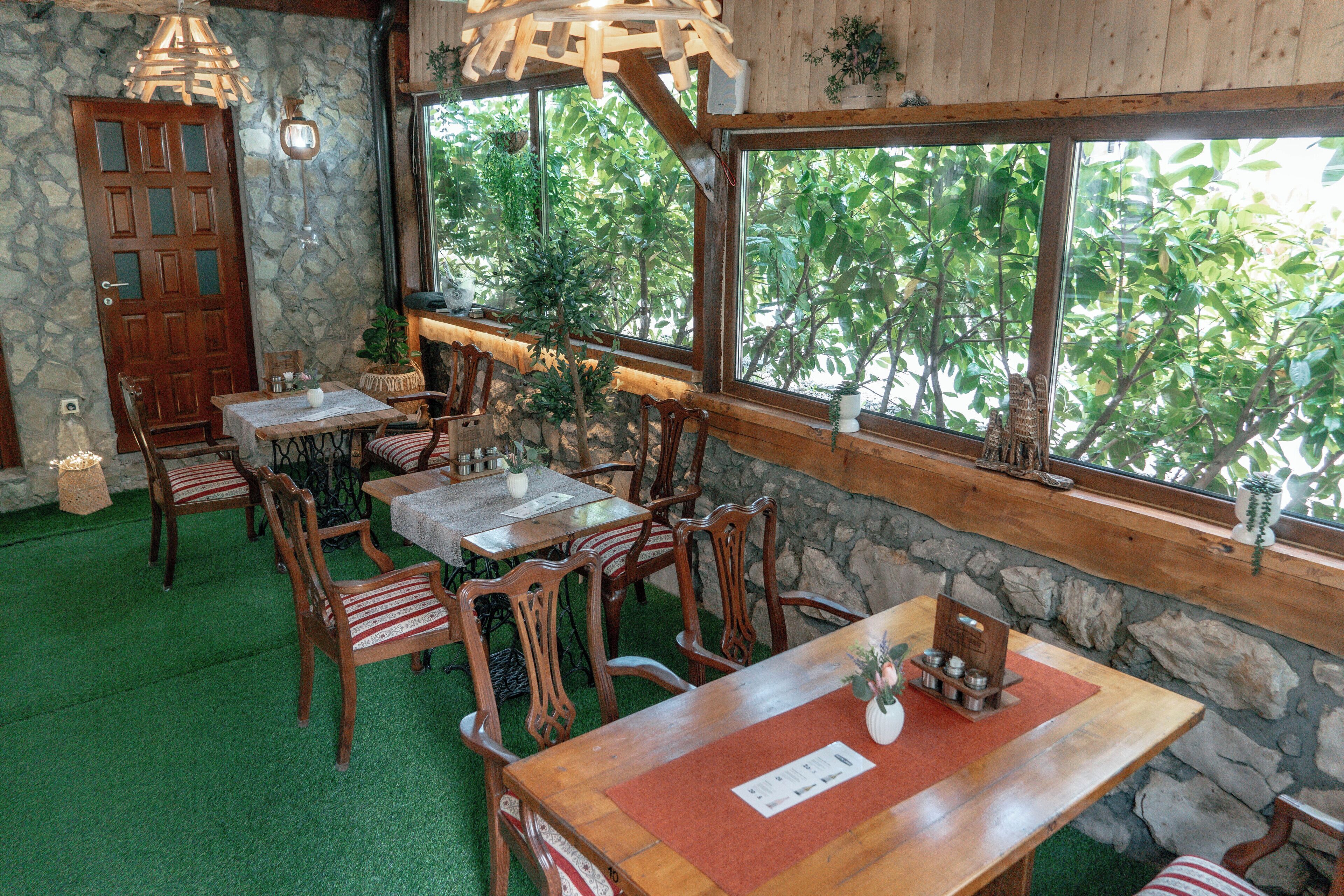 A restaurant with wooden tables and chairs, stone walls, and large windows overlooking greenery.