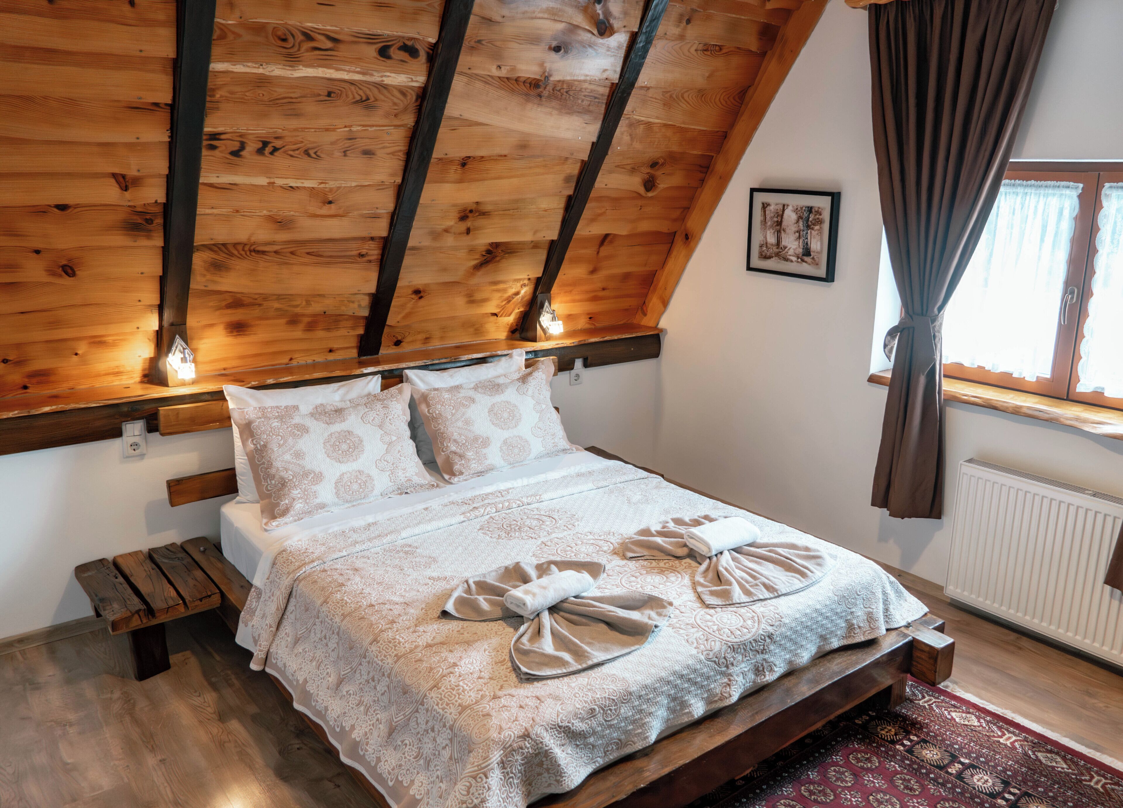 A bedroom with a wooden ceiling, a bed with patterned bedding, a nightstand, and a window with curtains.
