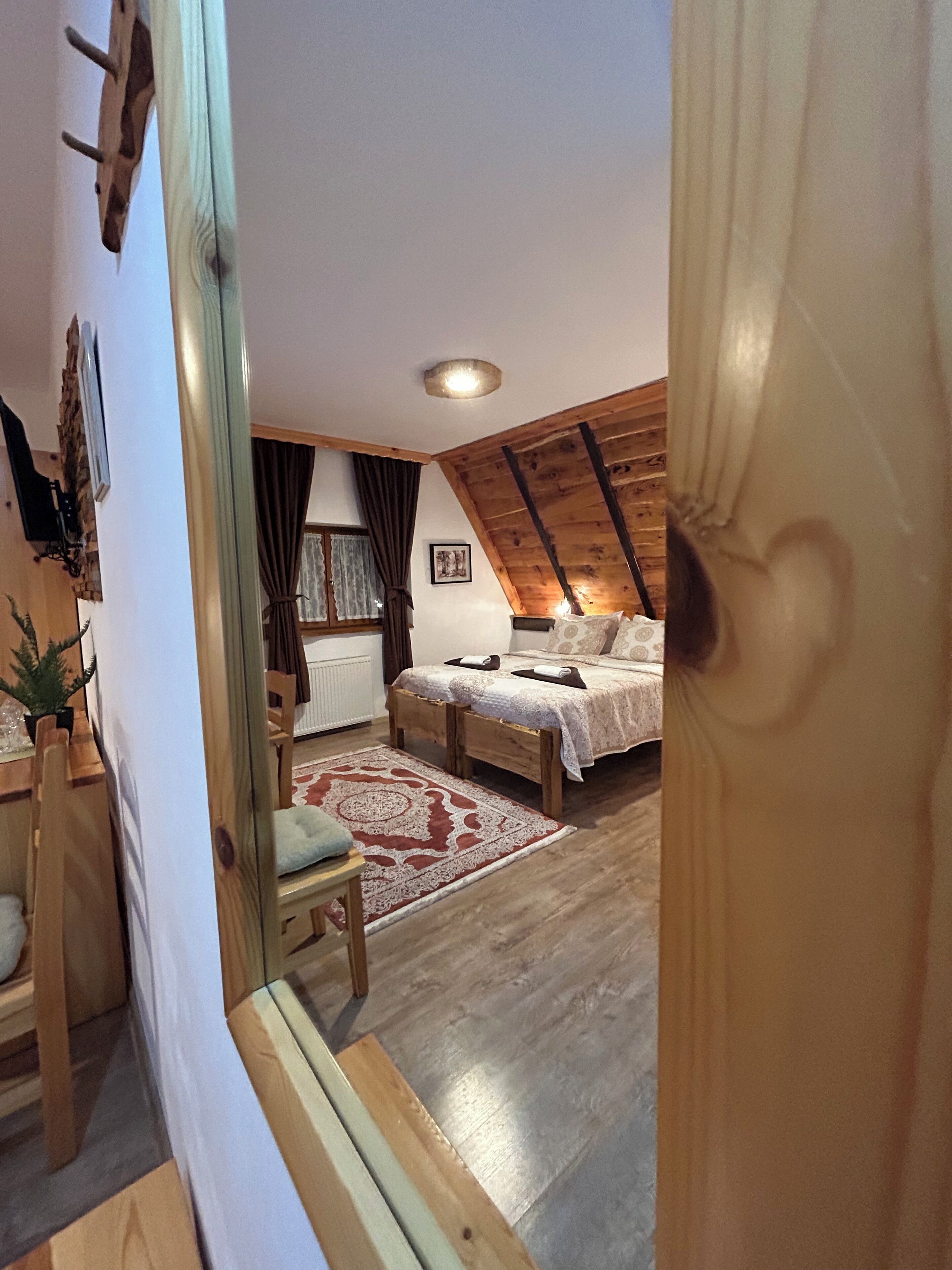 A bedroom with a wooden ceiling, a bed with a patterned bedspread, a bedside table, a chair, a small table with a plant, and a window with curtains.
