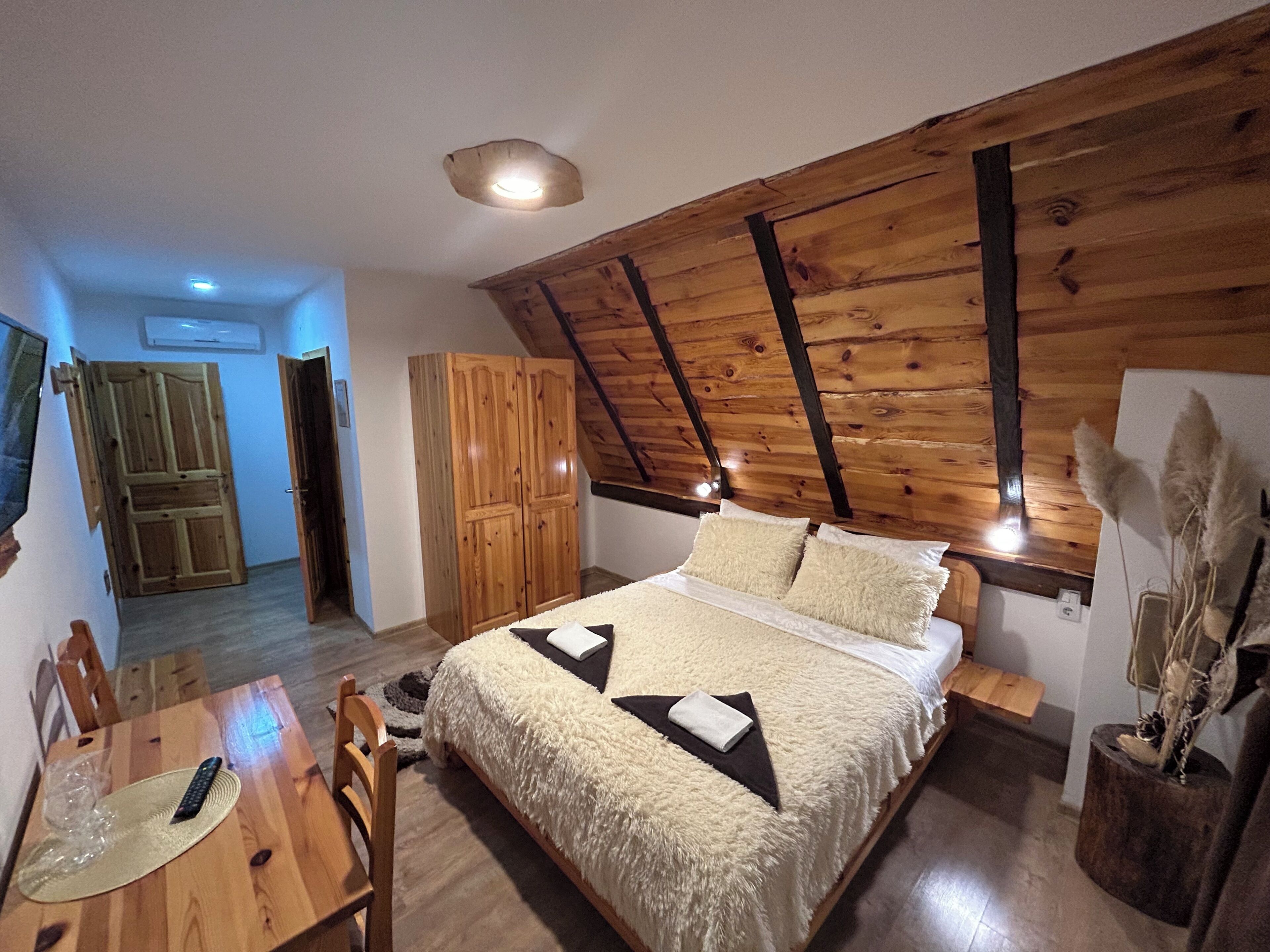 Une chambre avec un plafond en bois, un lit recouvert d’une couette moelleuse, une table en bois avec une télécommande, et une chaise.