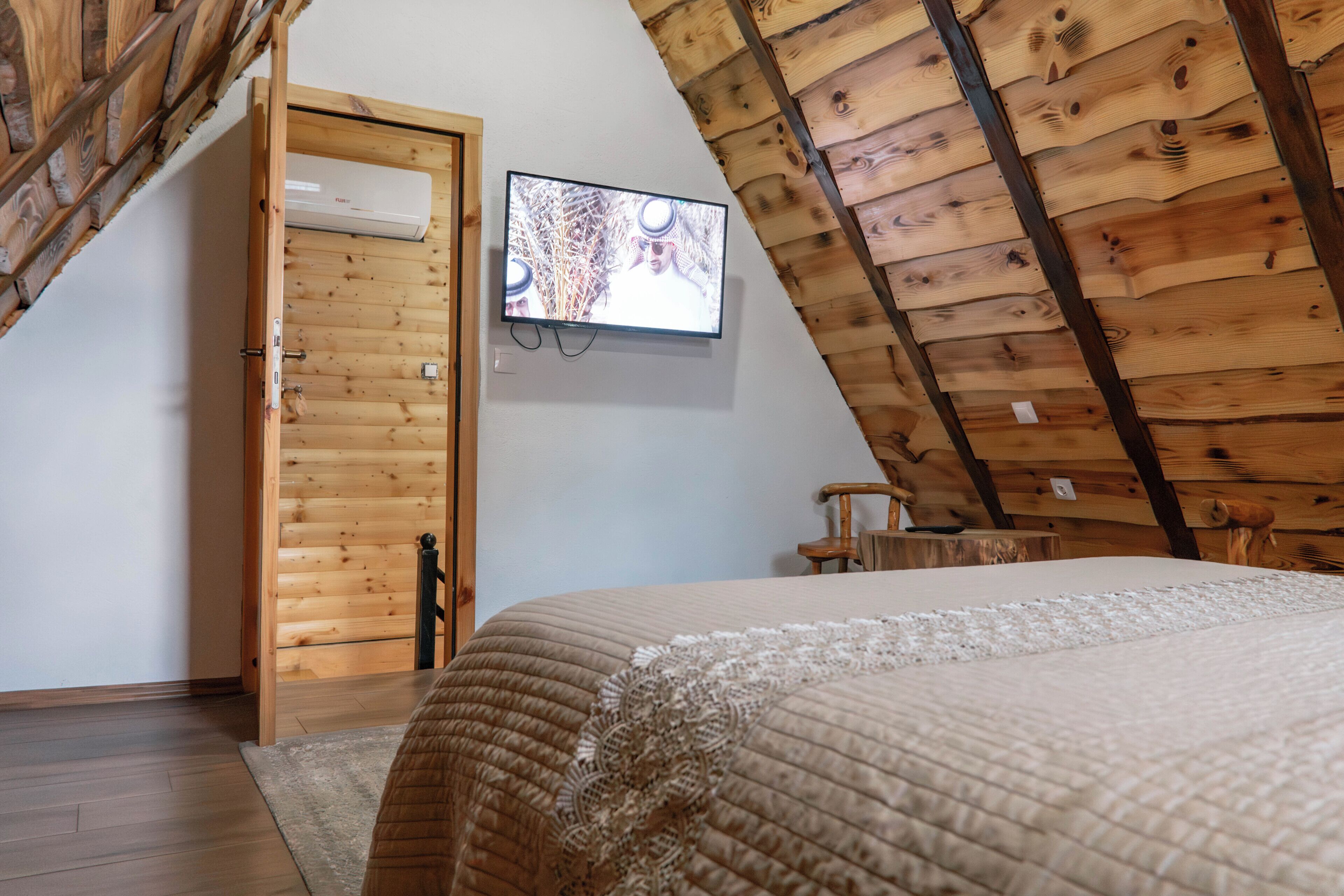 A bedroom with a sloped ceiling, a bed with a quilted cover, a television mounted on the wall, and a wooden door.