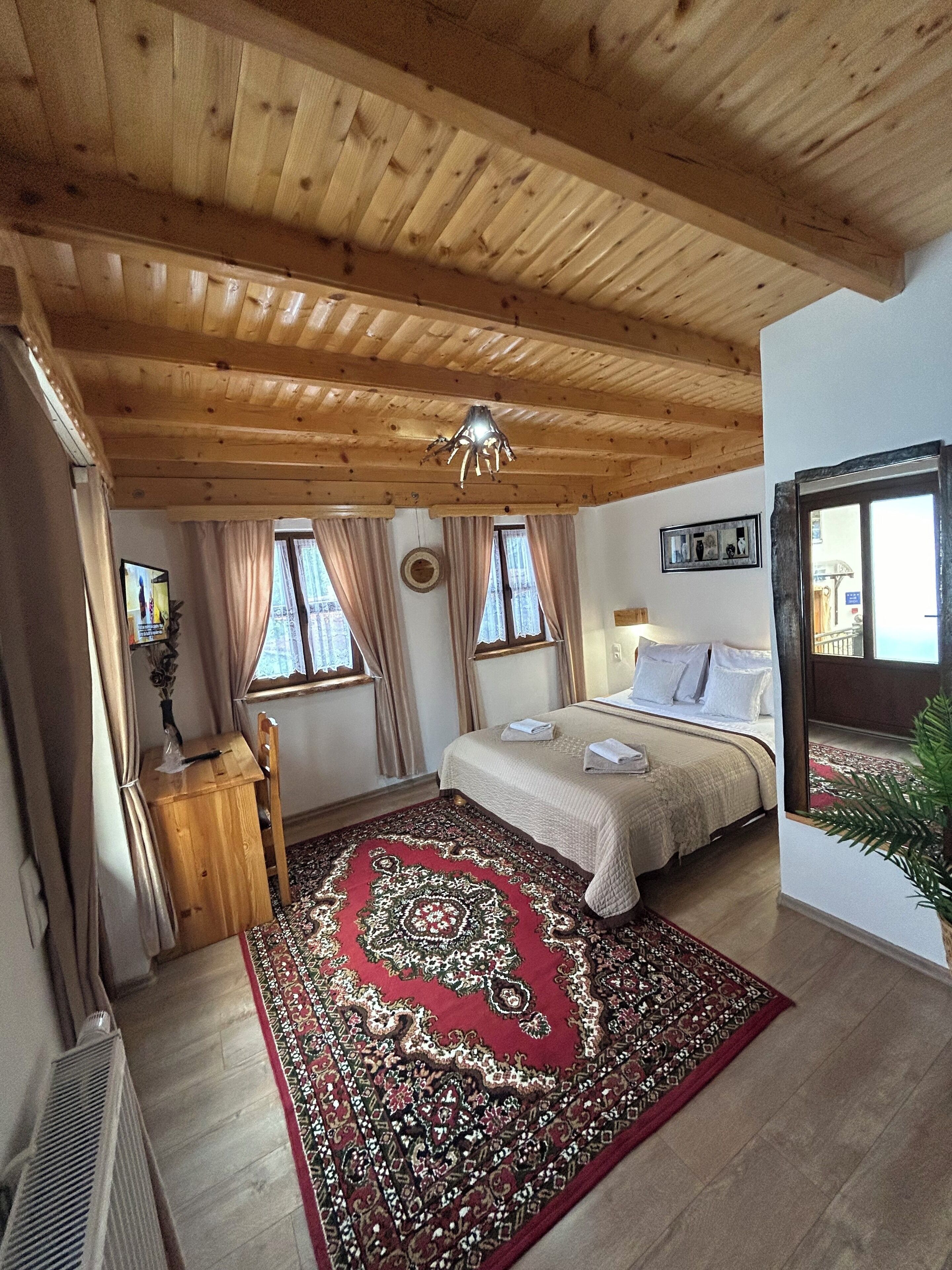 A bedroom with a wooden ceiling, a bed with white linens, a red patterned rug, and a small wooden desk with a chair.