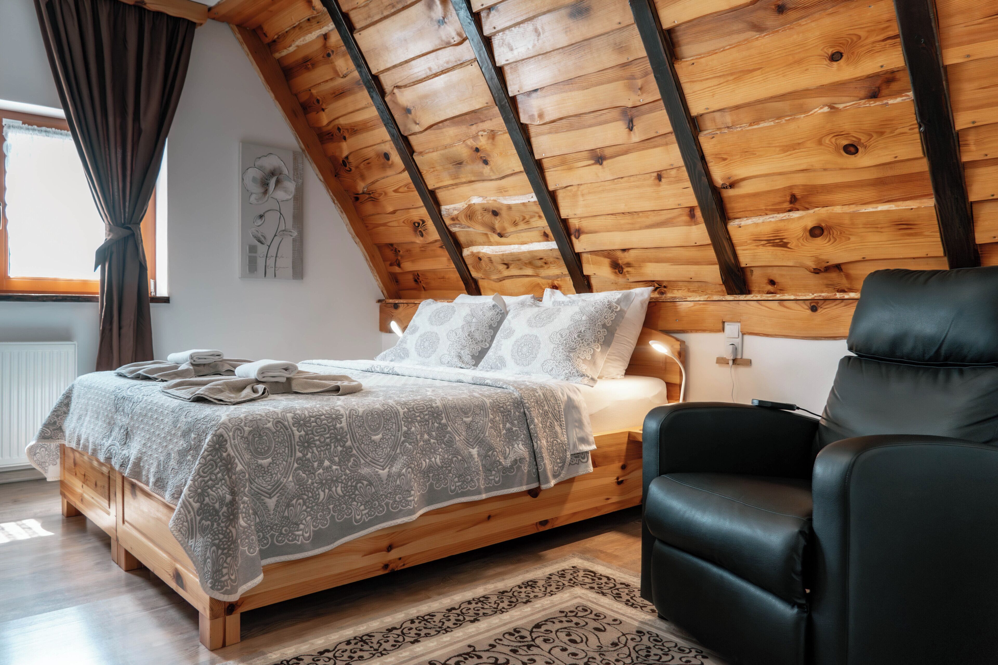 A bedroom with a wooden ceiling, a bed with patterned bedding, a black leather chair, and a window with curtains.