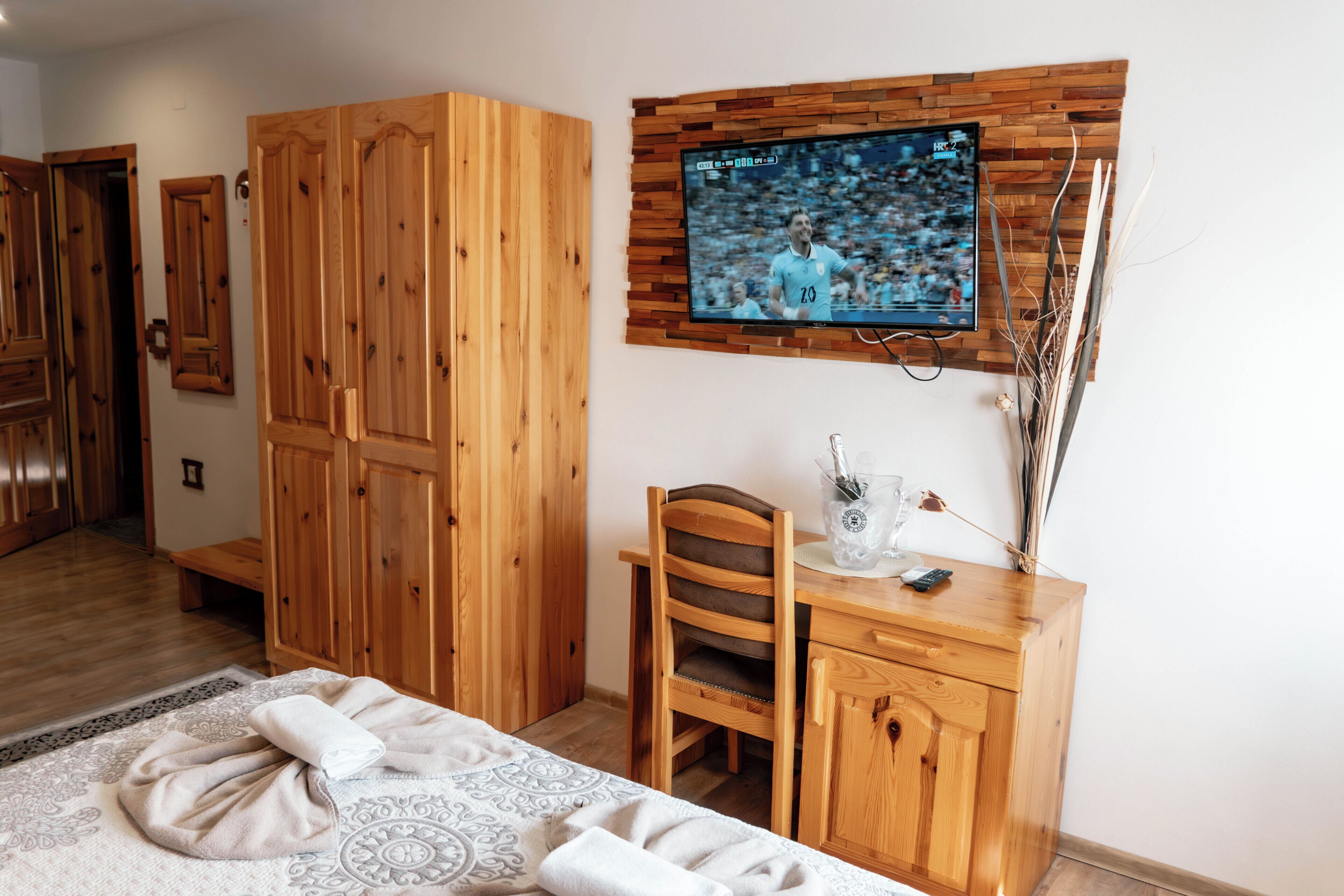 Une chambre comprenant un lit, une armoire en bois, un bureau avec une chaise, un téléviseur fixé au mur et un vase décoratif contenant des branches.