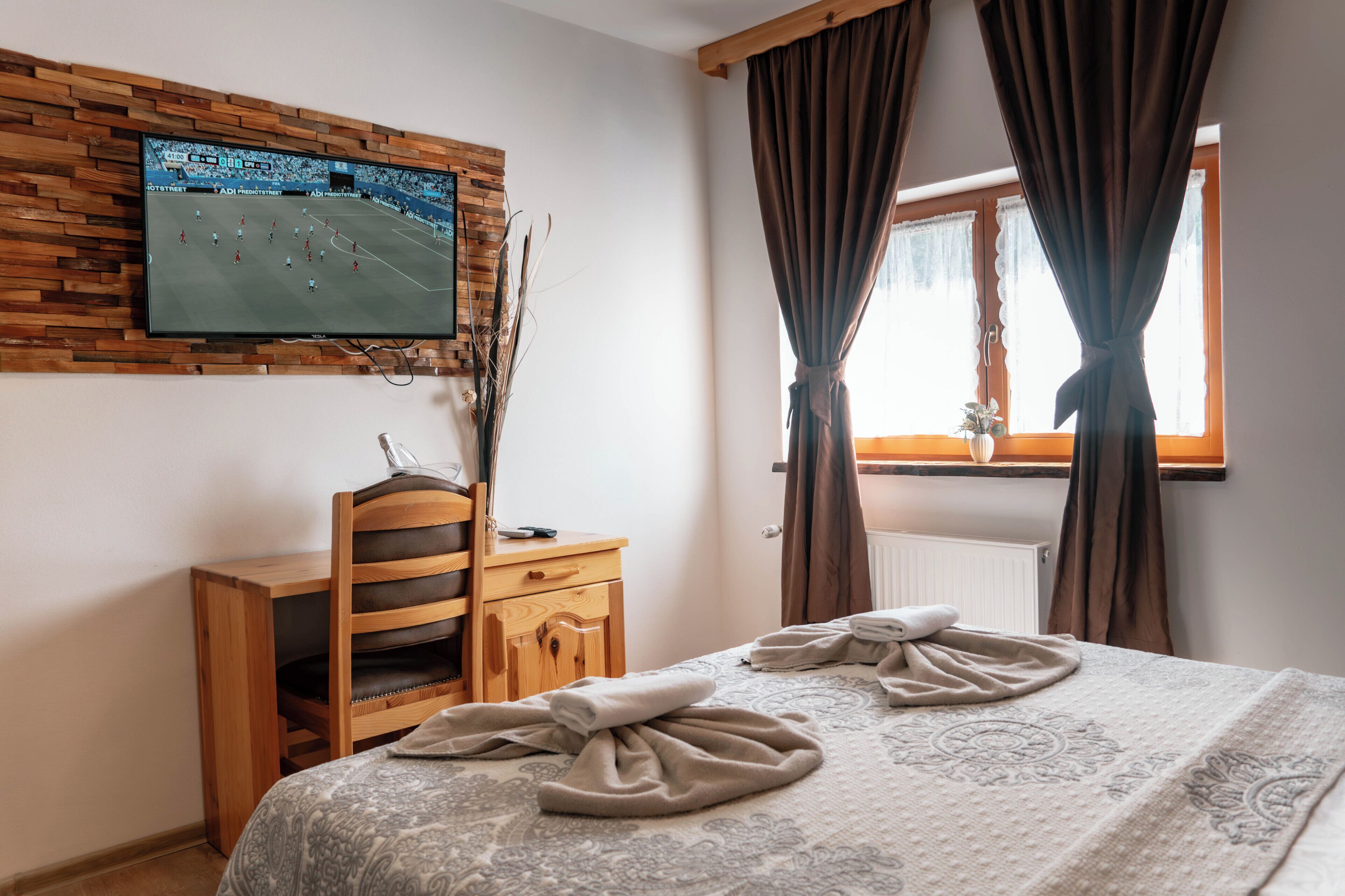 A hotel room with a bed, a desk, a television mounted on a wooden wall, and a window with curtains.