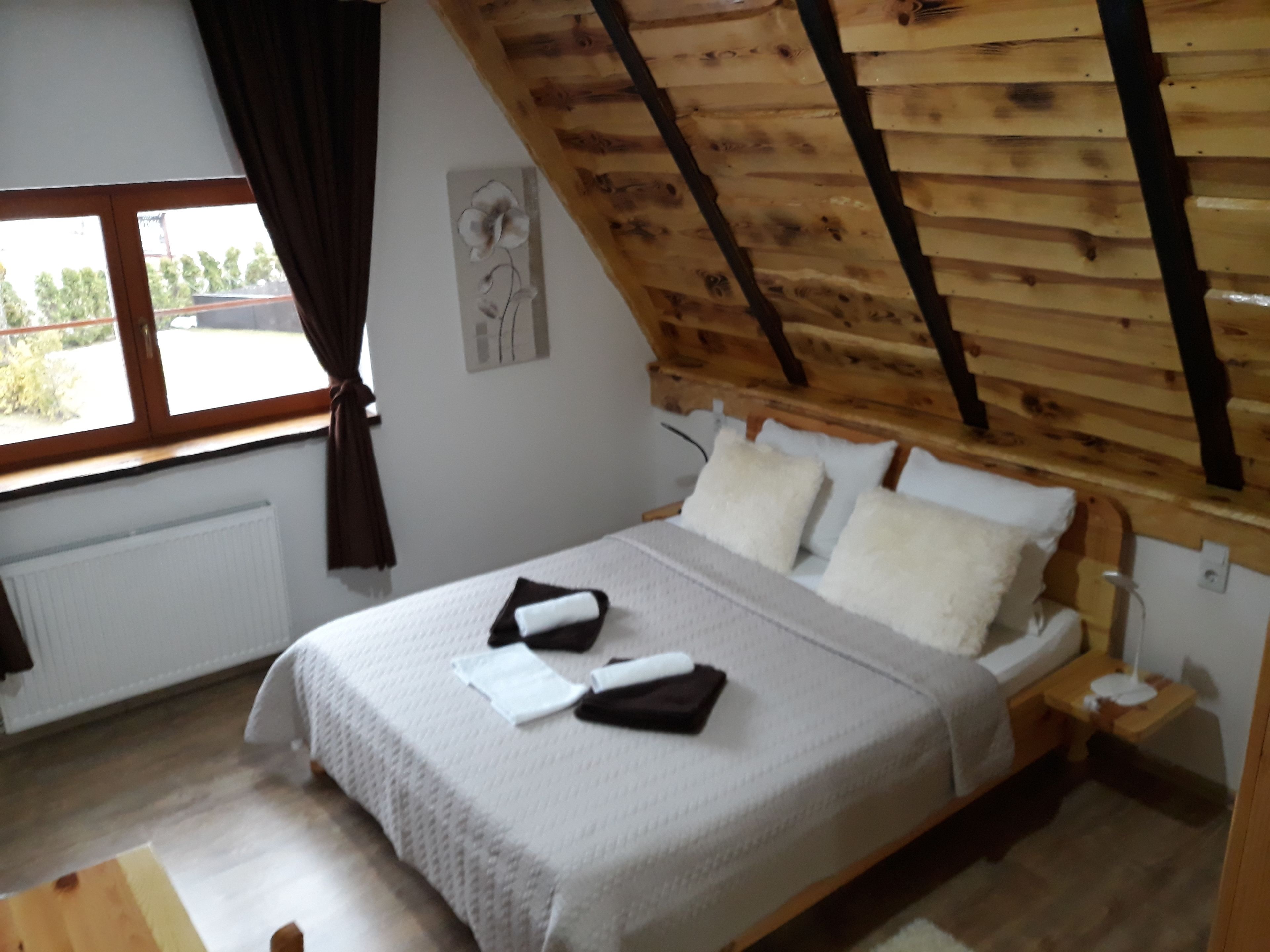 Un dormitorio con techo inclinado, una cama con ropa de cama blanca, una mesita de noche y un ventanal con cortinas.