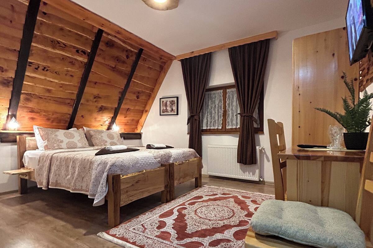 A cozy bedroom with a sloped ceiling, wooden furniture, a bed with a patterned bedspread, a desk with a chair, and a television.