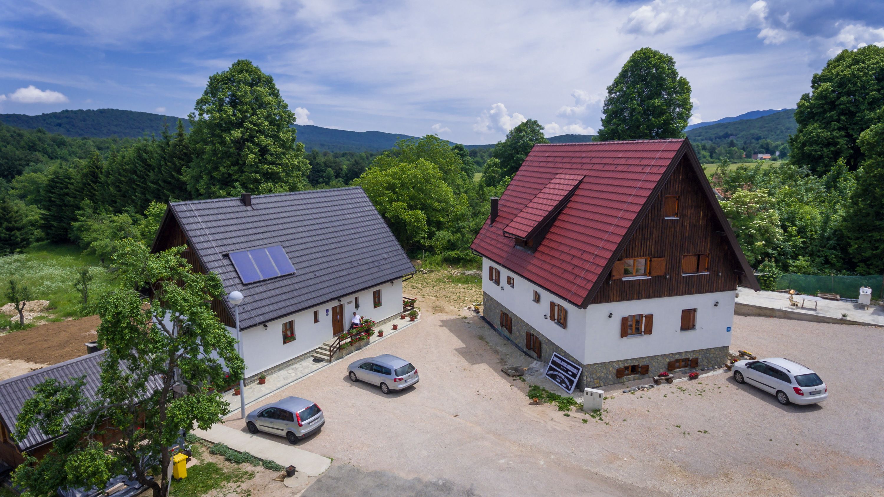 A rural setting with two traditional houses, one with a red roof and the other with a grey roof, surrounded by lush greenery and mountains in the background.
