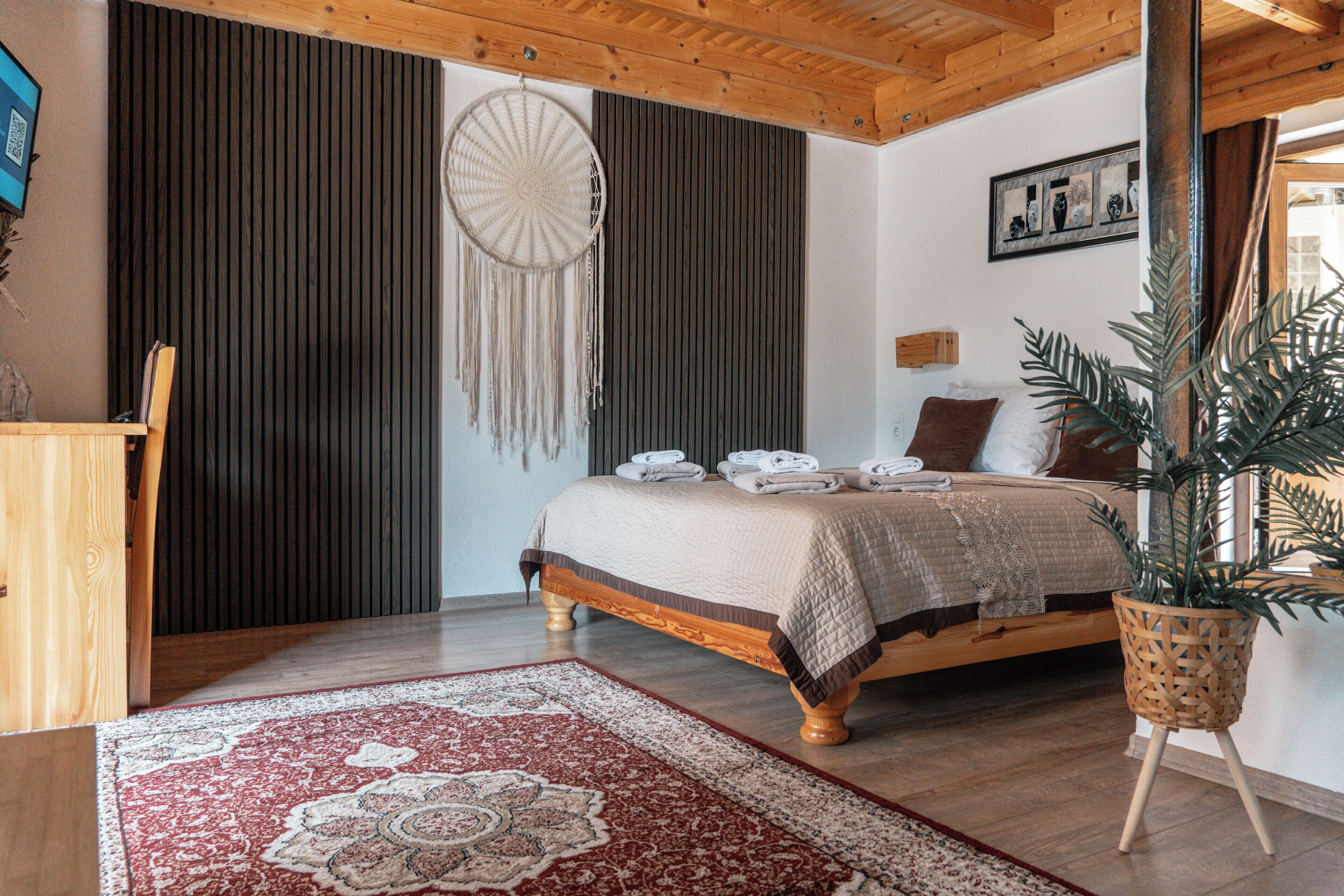 Une chambre avec un lit, une table de chevet, un attrape-rêves fixé au mur, une grande plante en pot et un plafond en bois.