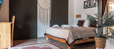 A bedroom with a bed, a bedside table, a wall-mounted dreamcatcher, a large potted plant, and a wooden ceiling.