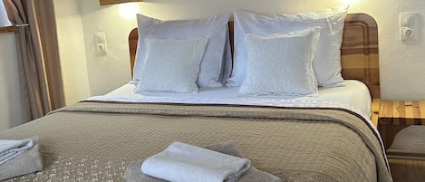 Una cama bien hecha con una colcha acolchada, dos almohadas y una toalla doblada. Encima de la cama hay una obra de arte enmarcada y un cabecero de madera.