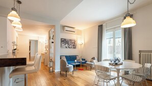 Un espacio habitable moderno con comedor, taburetes de bar, una mesa redonda con sillas de metal, un sofá azul y un amplio ventanal con cortinas.