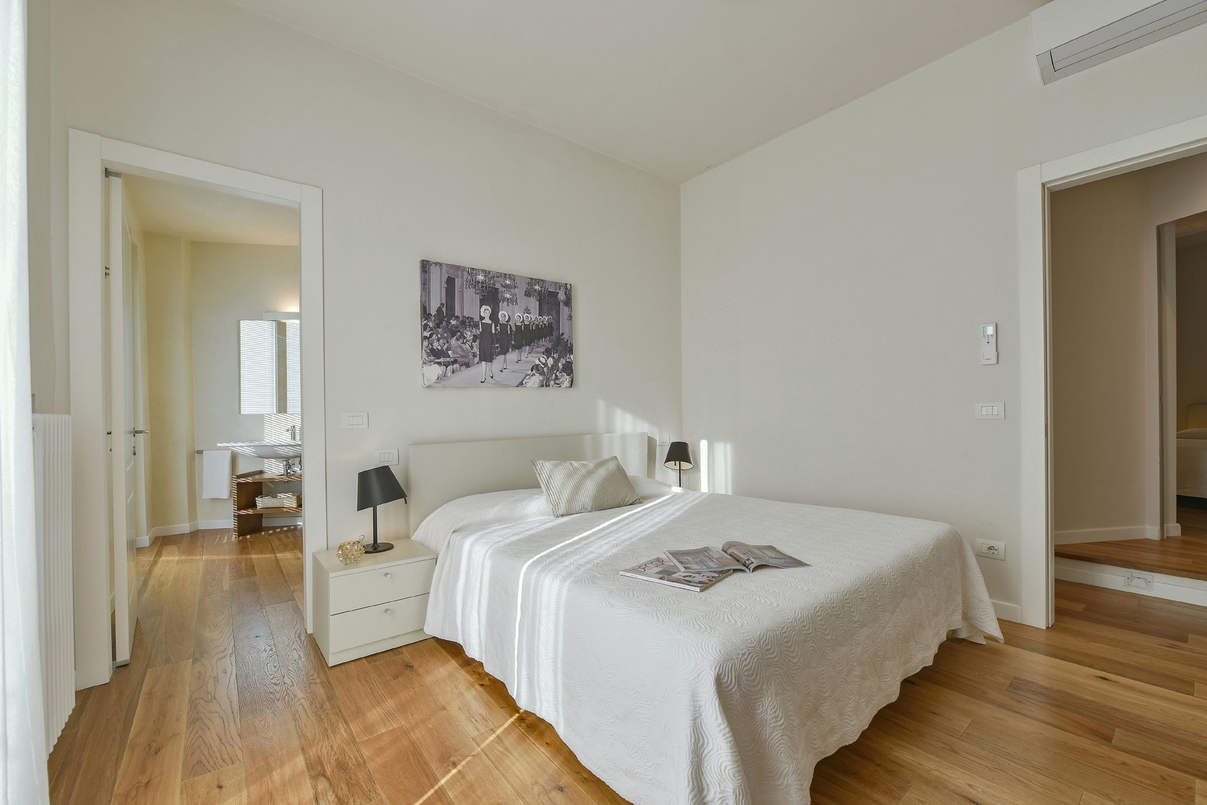 A bedroom with a bed, bedside tables, a lamp, a painting, and a wooden floor.