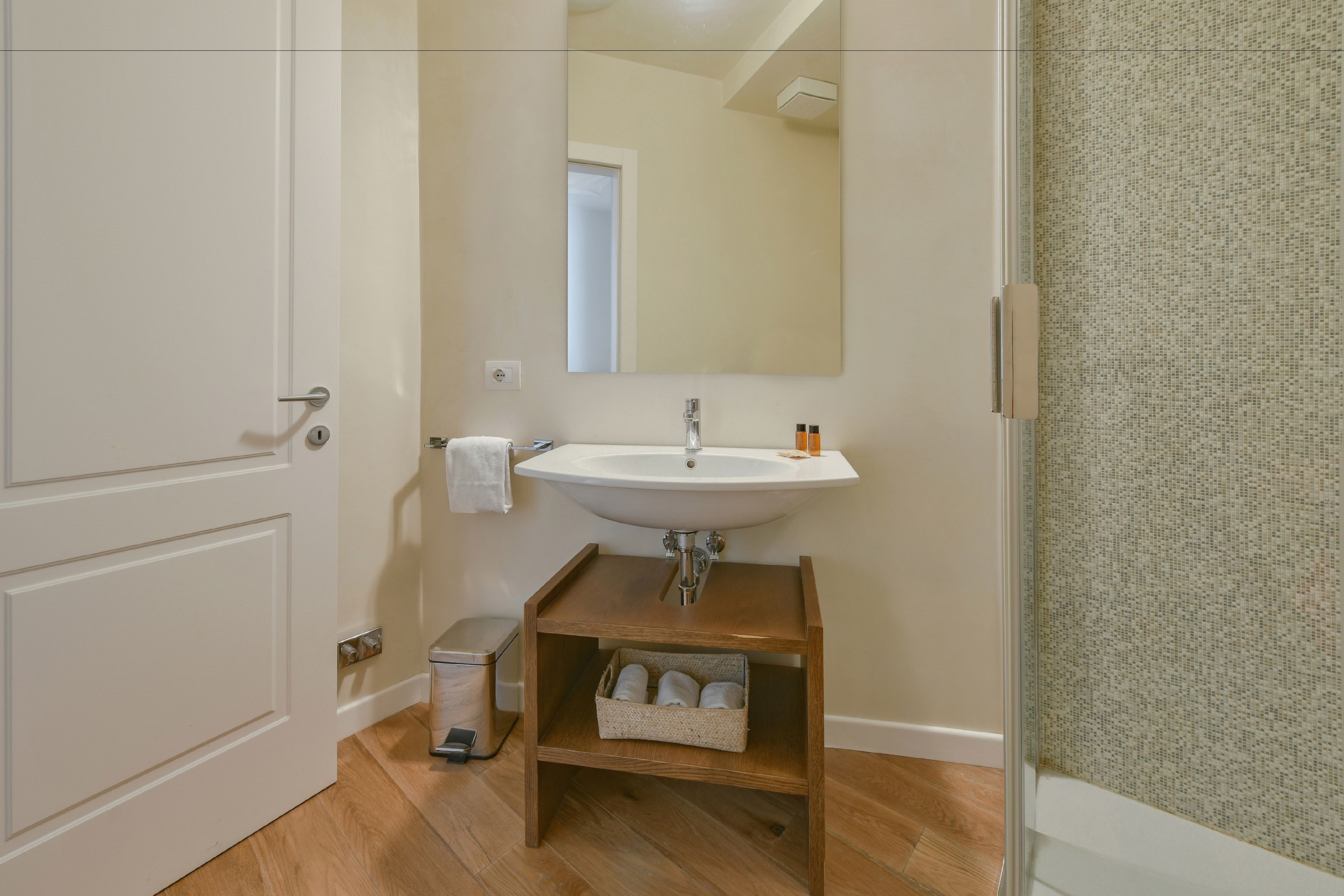 A bathroom with a white sink, a mirror, a towel rack, a small wooden shelf with towels, and a trash bin.