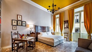 Une chambre d’hôtel avec un sol en pierre, un lit, un bureau avec des chaises, un lustre et des tableaux encadrés accrochés au mur.
