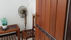 Uma porta de madeira com fecho de metal, uma televisão presa à parede, um ventilador de chão, uma pequena mesa de madeira com um vaso de planta e uma cadeira de madeira.