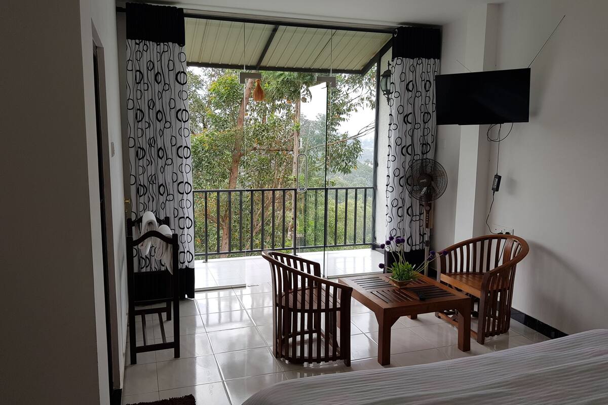 A room with a bed, a table, and two chairs. There is a balcony with a view of trees and a railing.