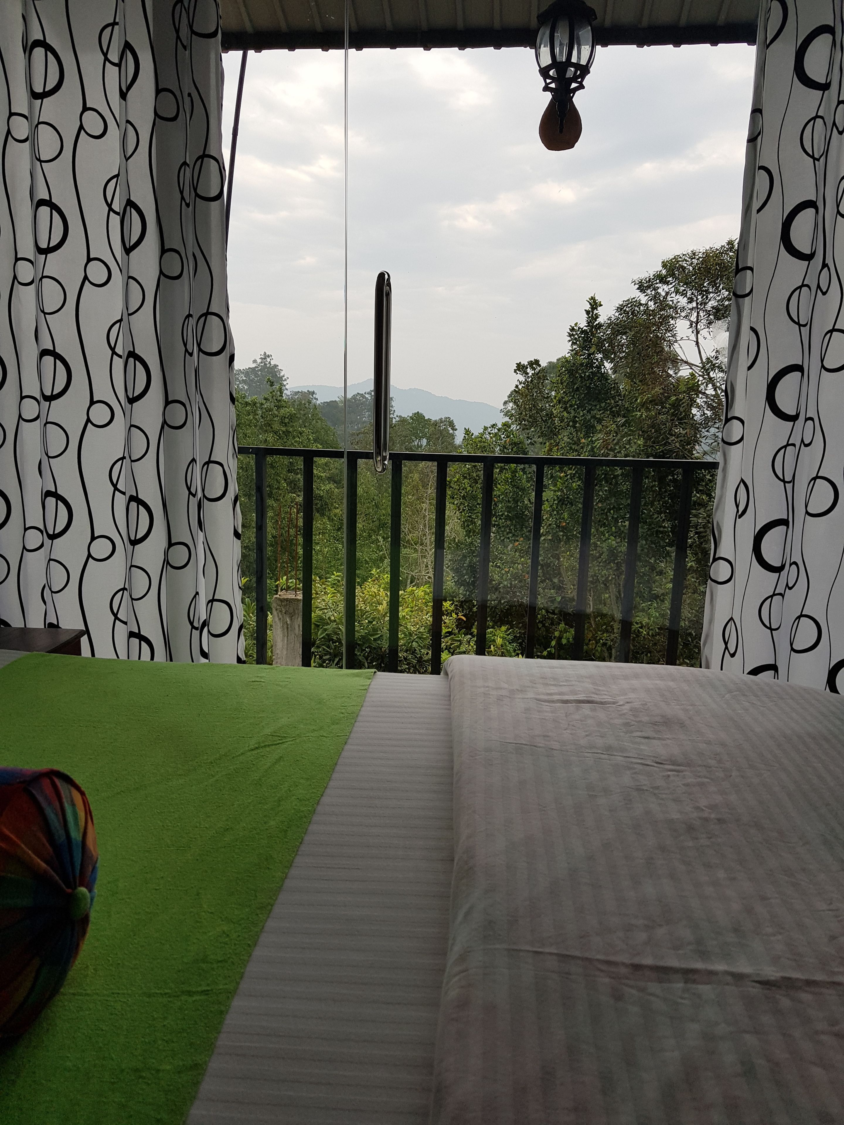 Un balcon avec vue sur les arbres et les montagnes, un luminaire suspendu et un lit avec une literie aux tons verts et gris.