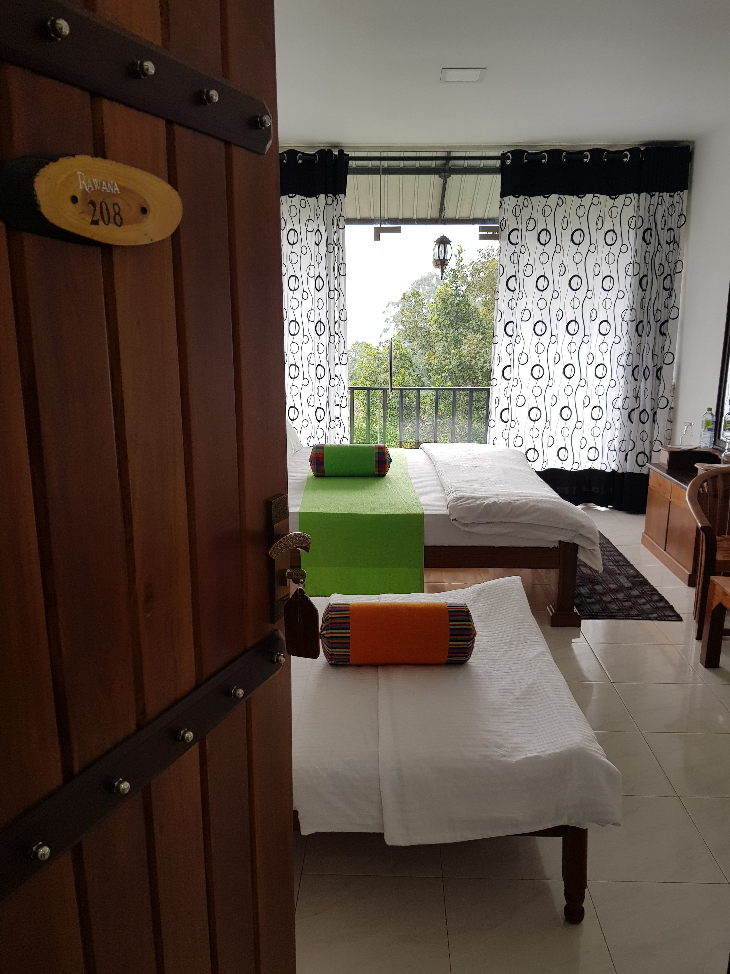 A hotel room with a wooden door, a bed with a green pillow, a bedside table, and a view of the outdoors through patterned curtains.