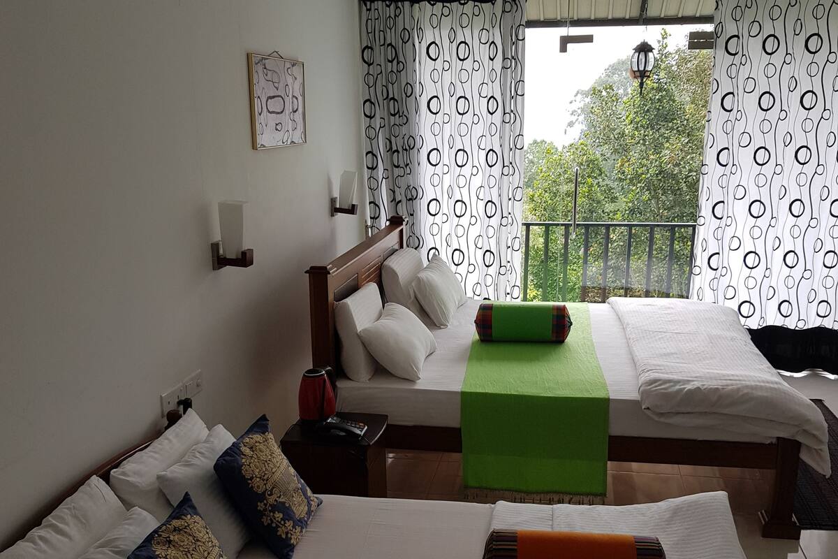 A hotel room with two beds, a green bed runner, and a view of greenery outside.