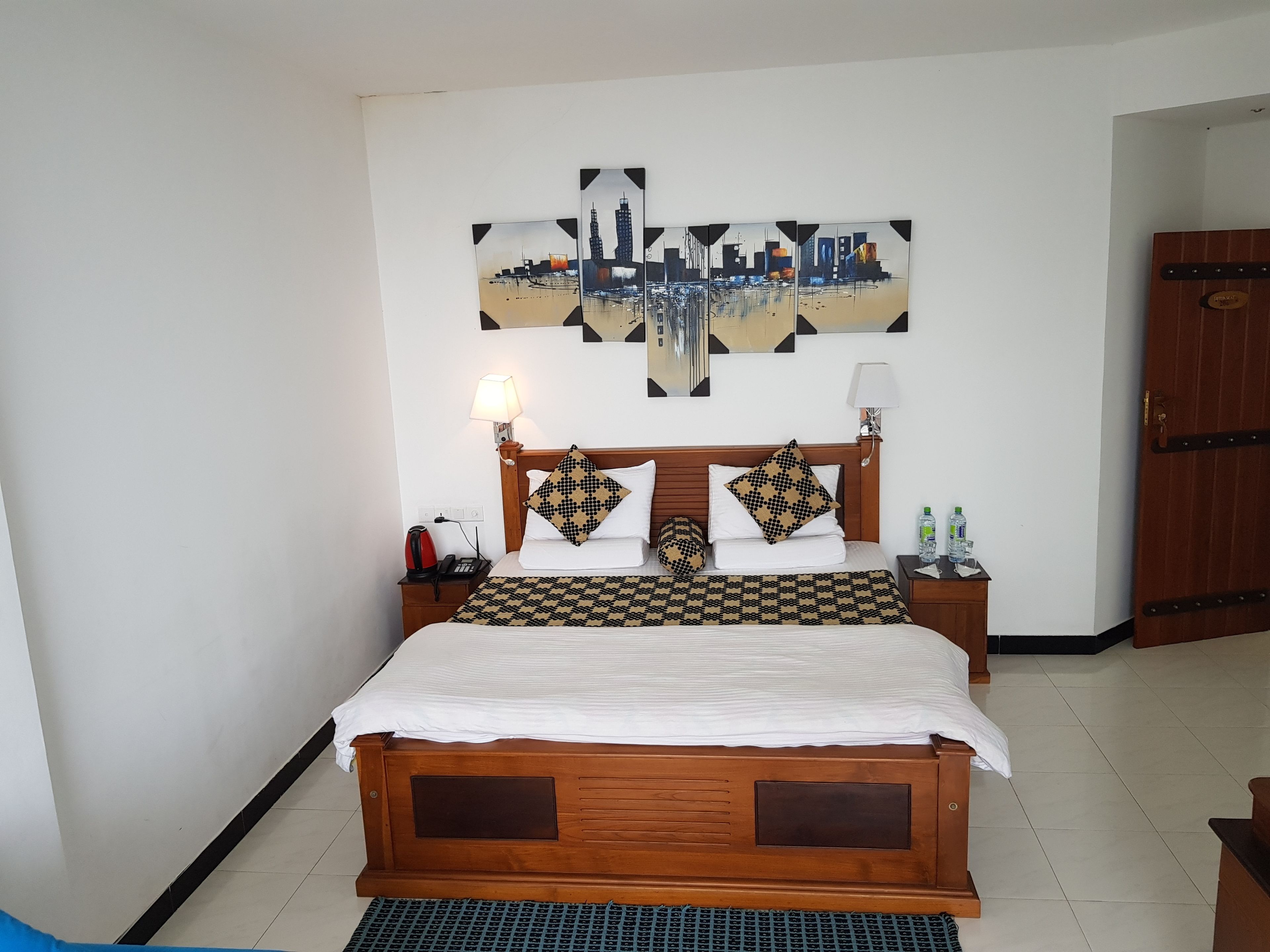 A neatly made bed with a patterned bedspread, two bedside lamps, a nightstand with a kettle, and a wall-mounted cityscape artwork.