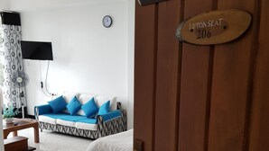 Quarto de hotel com cama, sofá de padrão azul e branco, mesinha, relógio na parede e porta de madeira com placa de número de assento.