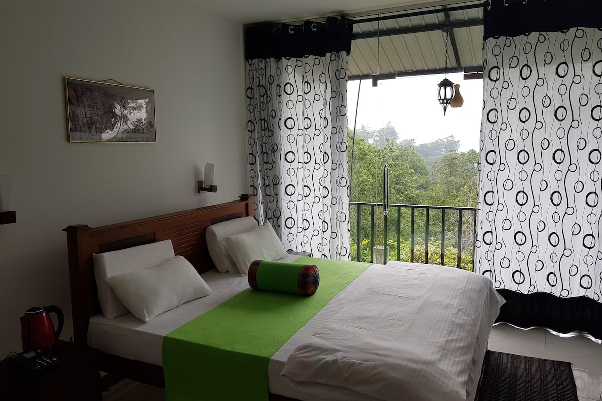 A hotel room with a bed, a green throw blanket, a painting on the wall, and a view of the outdoors.
