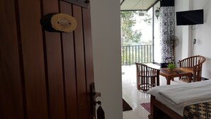 Quarto com porta de madeira, cama, mesa com cadeiras, TV fixada na parede e varanda com vista para árvores.