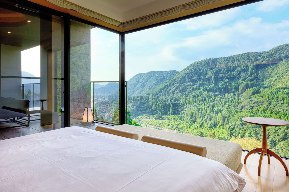 Une chambre avec un grand lit, une petite table ronde et une vue sur une chaîne de montagnes boisées.