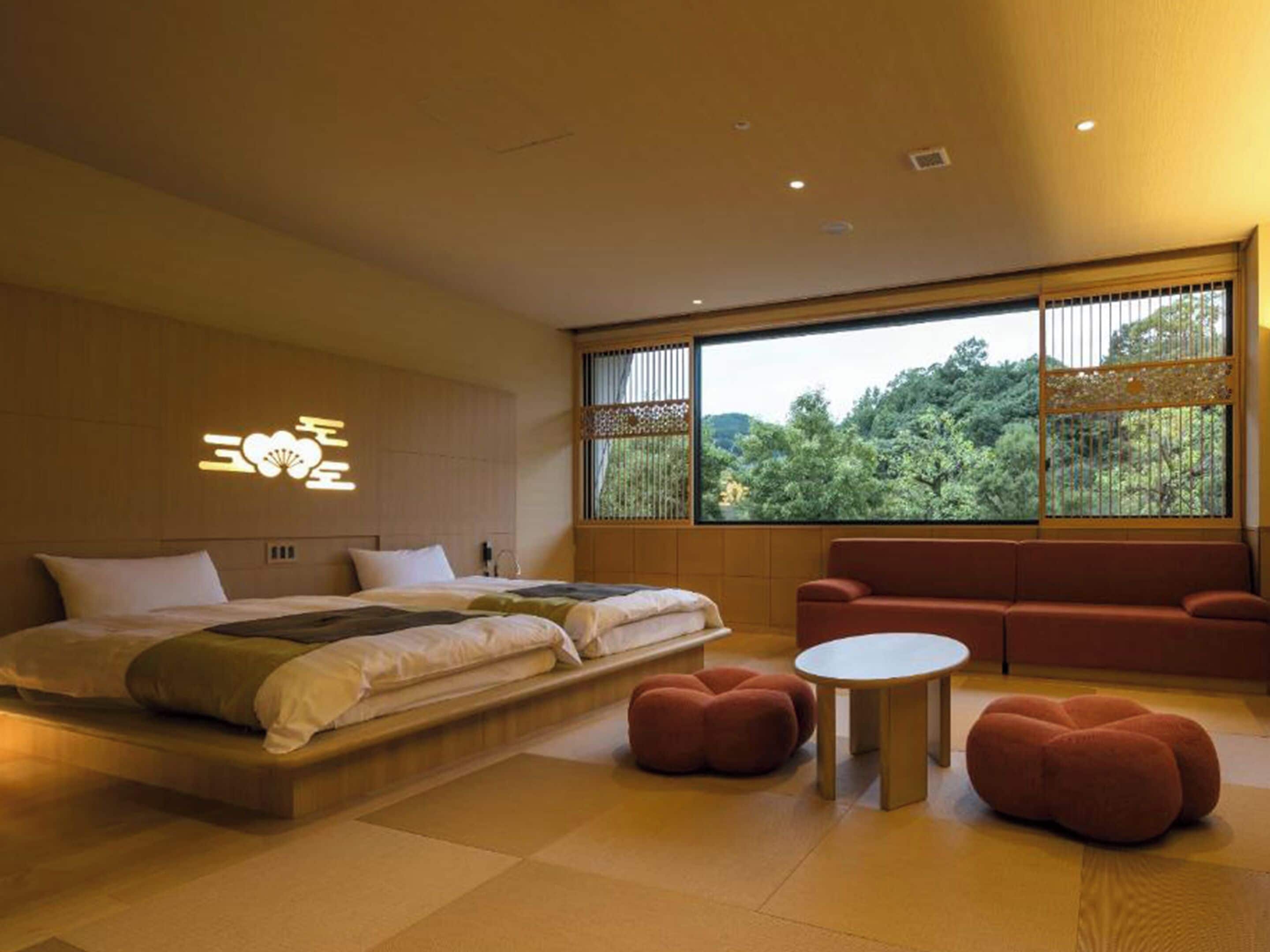 Une chambre moderne avec un grand lit, un canapé, une petite table et une vue sur la verdure grâce à de grandes fenêtres.