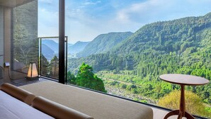A bedroom with a large window offering a view of a forested valley and mountains.