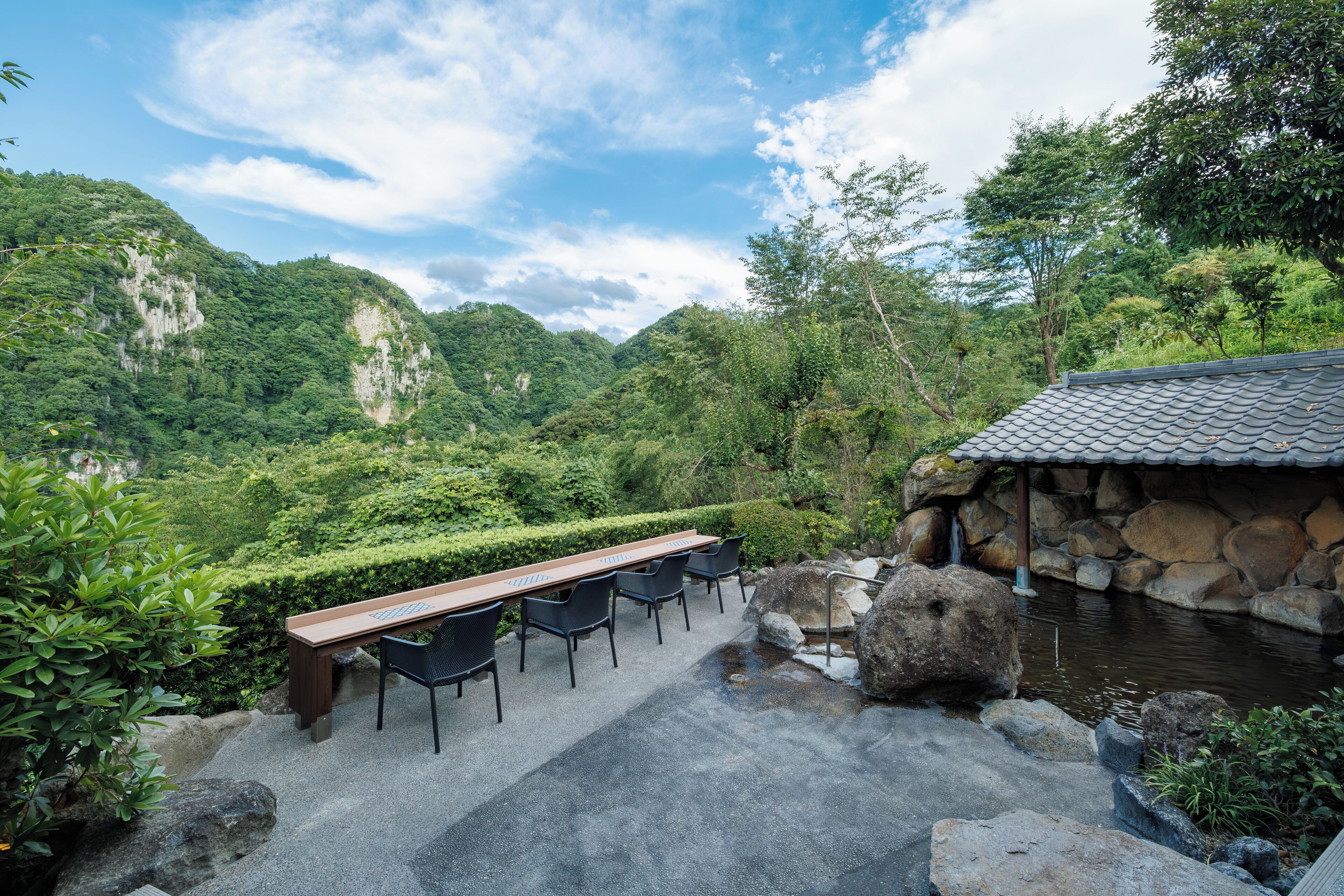 A serene outdoor seating area with a long wooden table and chairs, surrounded by lush greenery and a small pond.