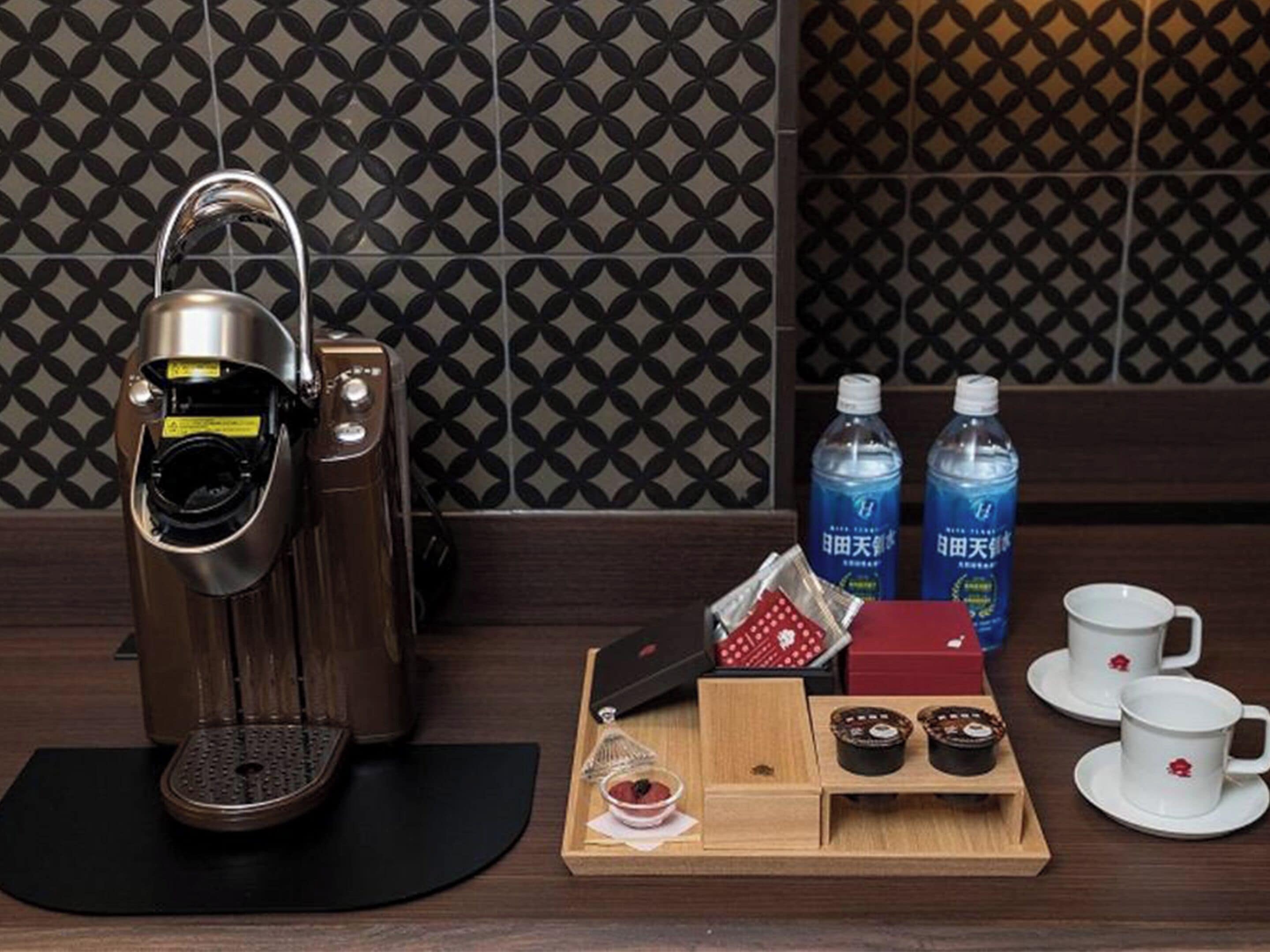 A coffee machine, two bottles of water, a tray with snacks, and two cups on a wooden table.