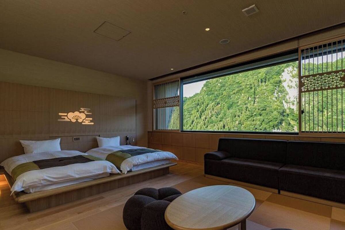 Une chambre d’hôtel moderne, dotée d’un grand lit, d’un canapé, d’une petite table ronde et offrant une vue sur une colline boisée.