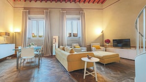 Un salon spacieux comprenant un canapé, un pouf et un coin repas avec des chaises et une table. On y trouve également un téléviseur et une lampe.