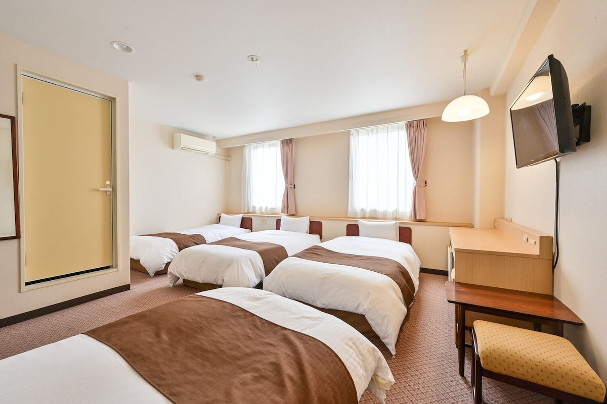 A hotel room with four single beds, a desk, a chair, a television, and a window with curtains.