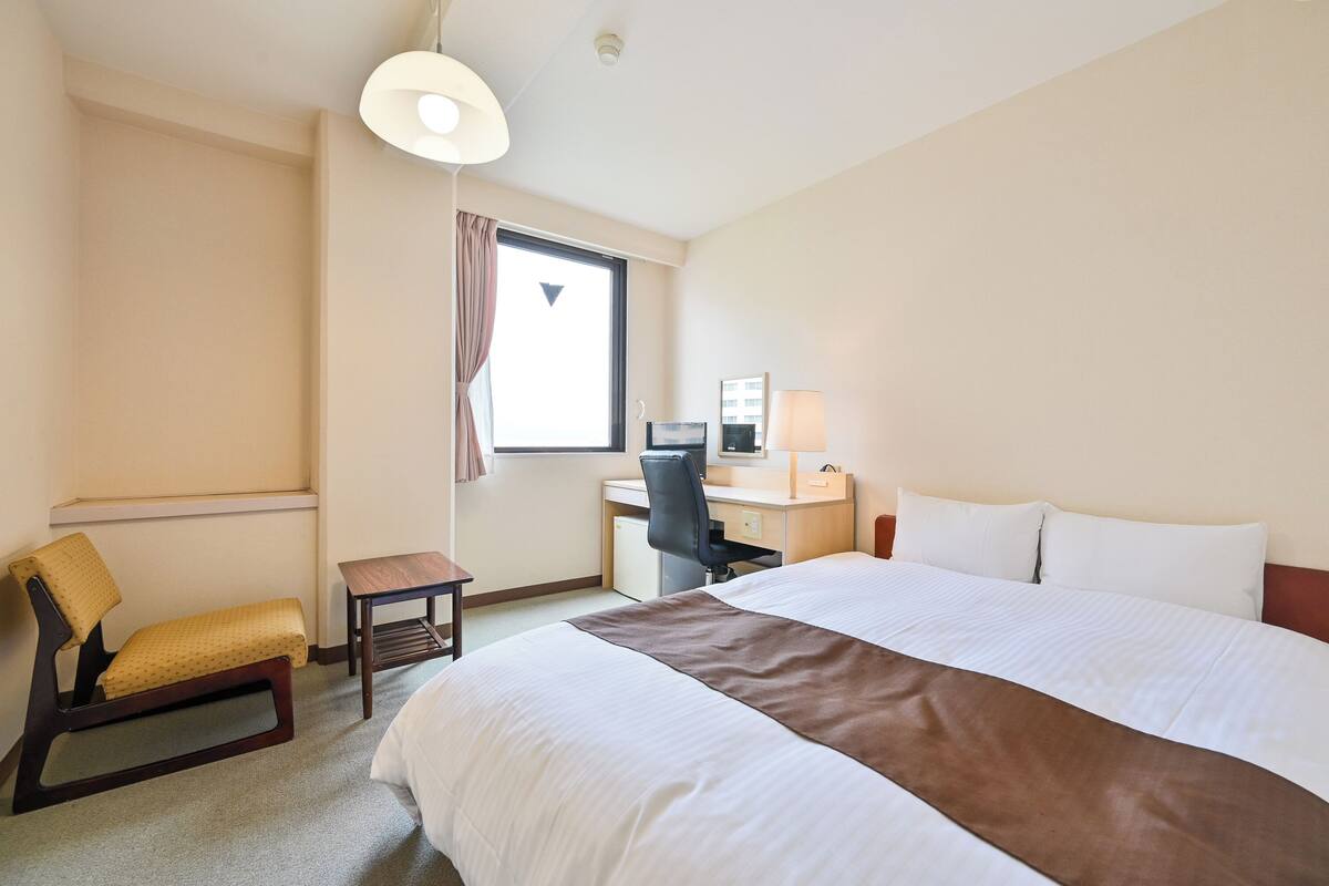 A hotel room with a large bed, a desk with a chair, a small table, and a window with curtains.